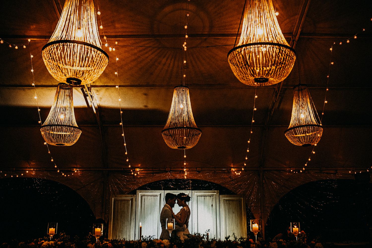 Romantic silhouette of couple kissing under elegant chandelier lights at wedding reception.