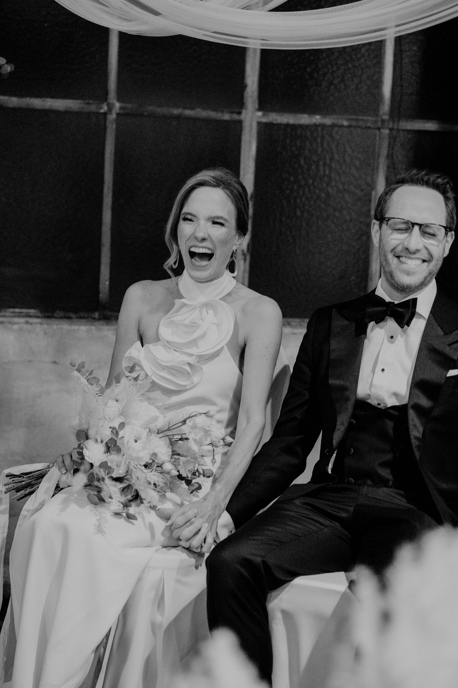 A black and white wedding photo of the bride in a white gown and groom in tuxedo laughing together during reception.