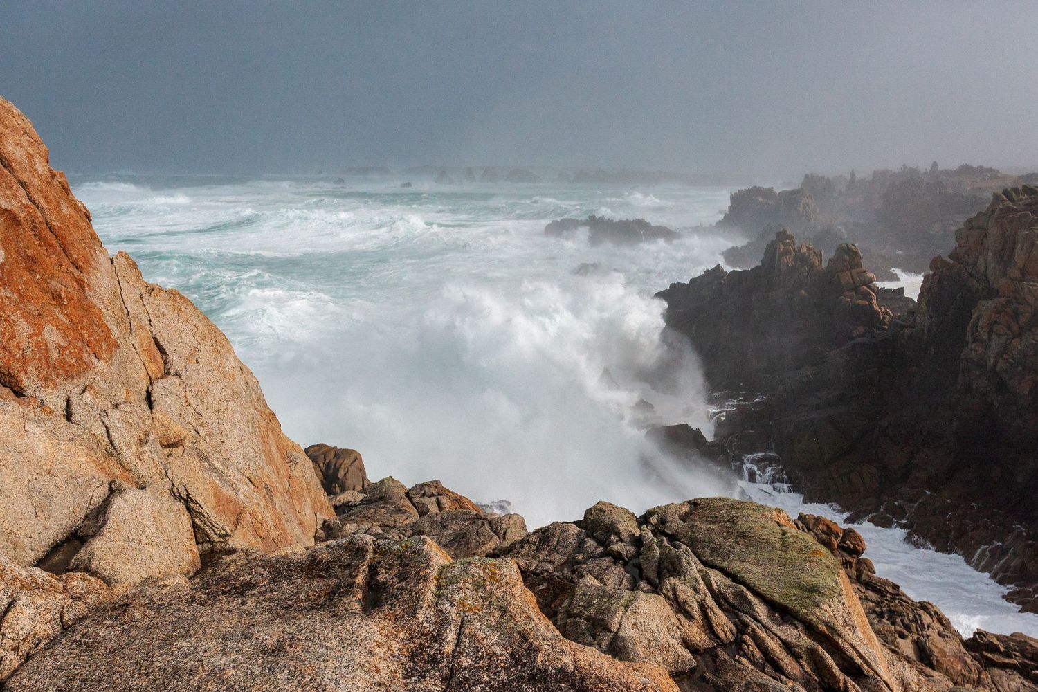 Powerful ocean waves smash into rocky coastline under stormy grey skies.