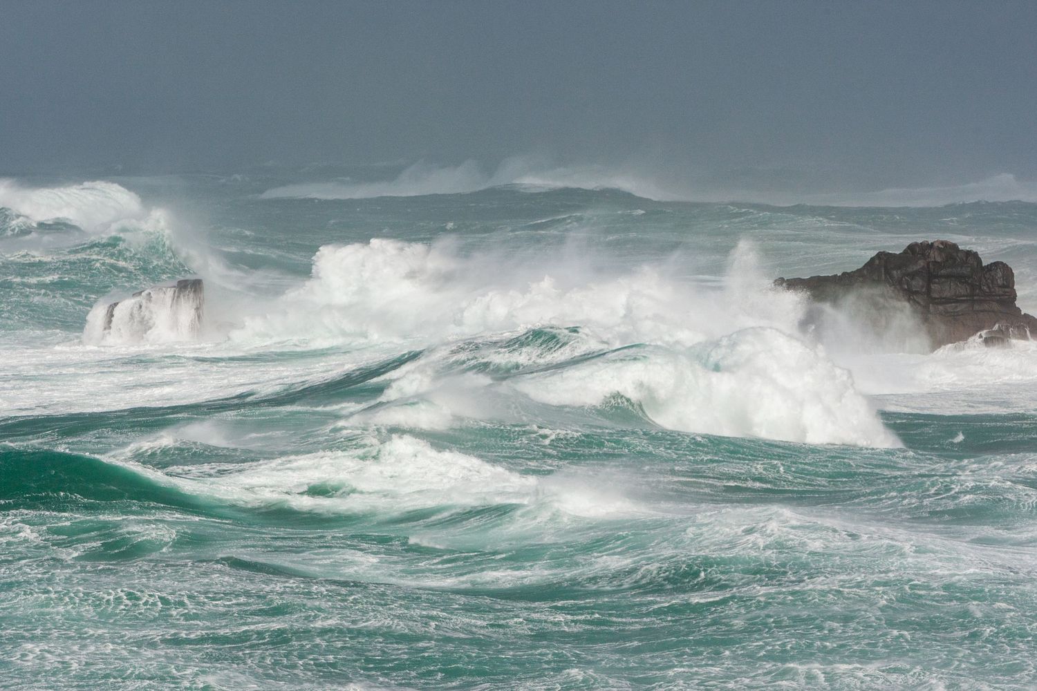 Turquoise storm waves crash and spray over rocky coastal outcrops.