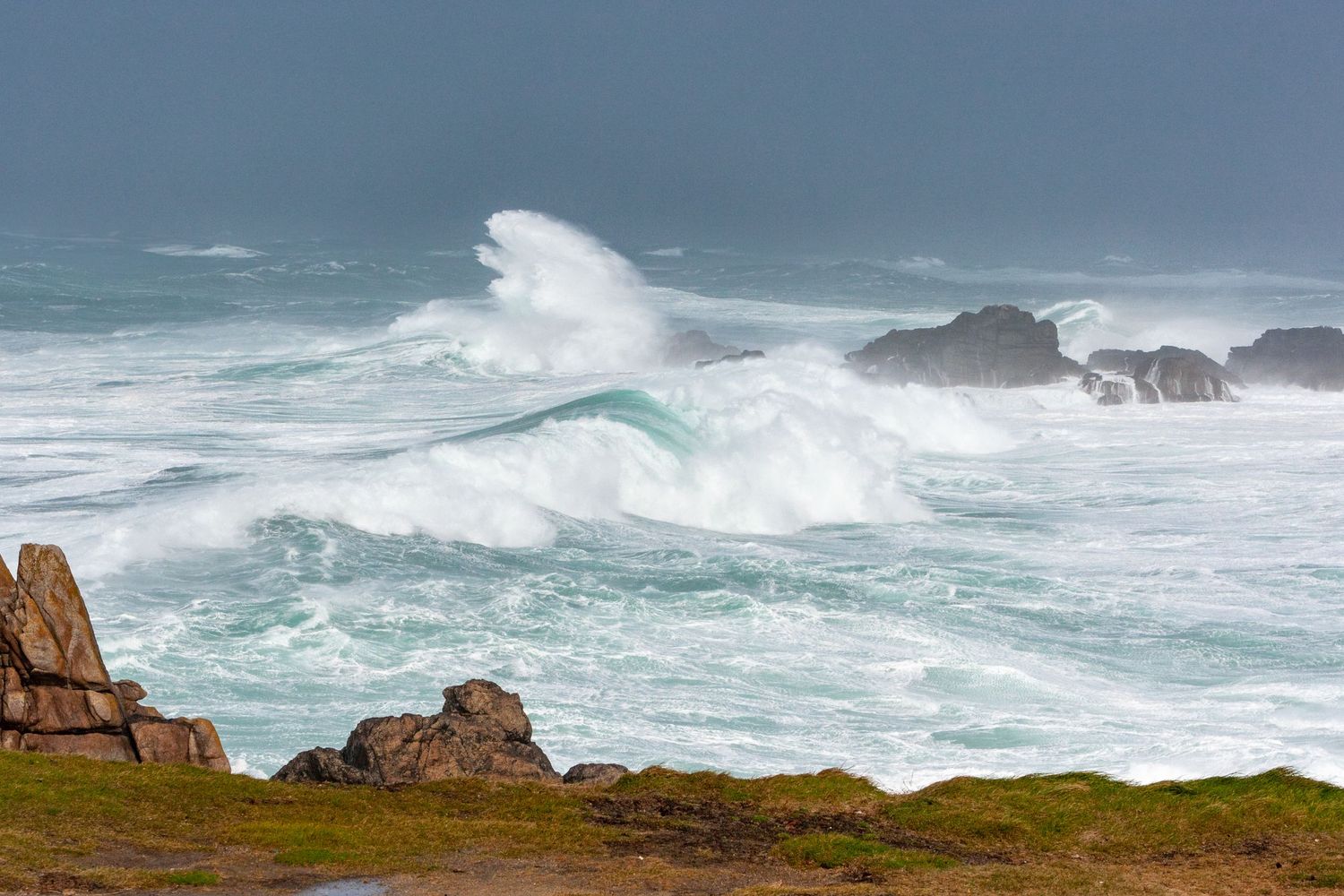 Giant waves break over rocky shoreline with grass-covered cliffs in foreground.