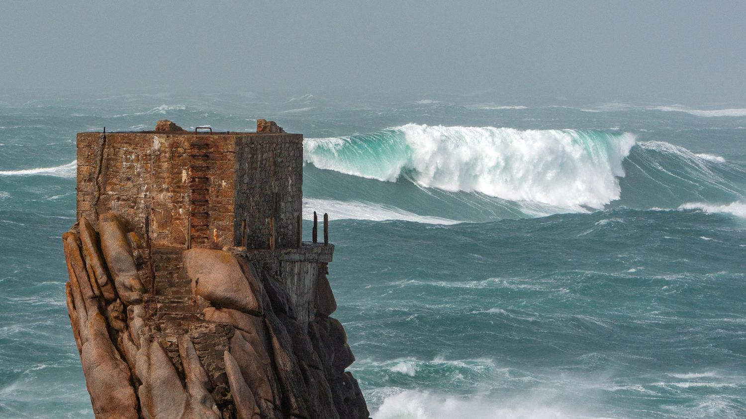 Historic stone fortress perched on cliff edge faces massive ocean waves.