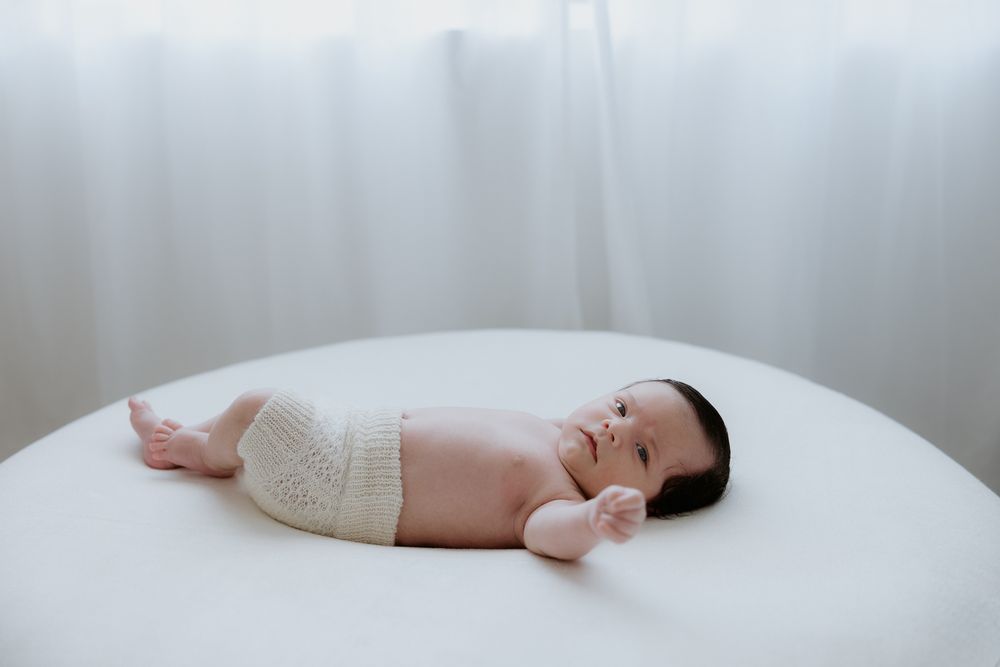 Newborn baby sleeps peacefully on soft white bedding while wearing a white onesie.