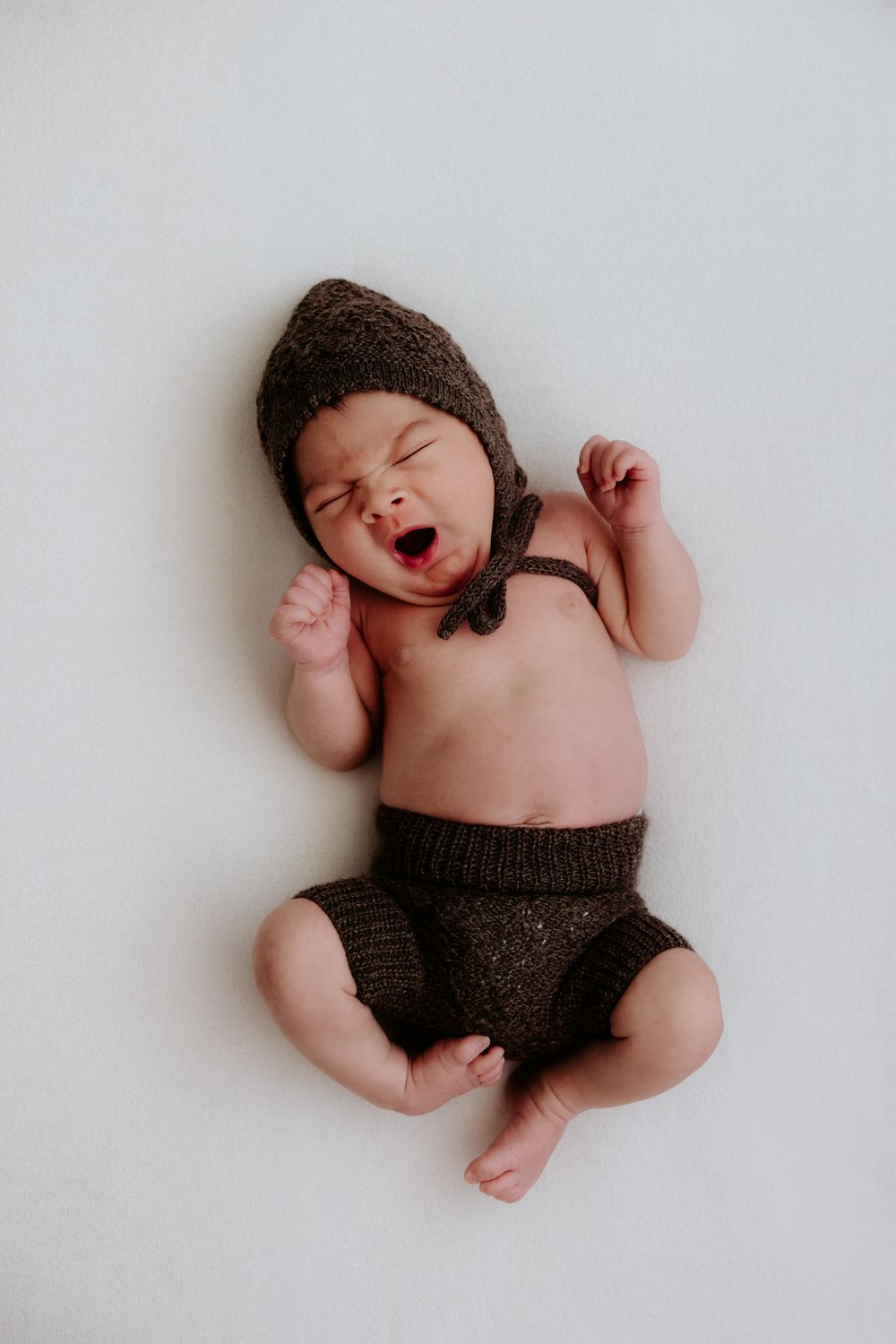 Newborn baby wearing brown knit hat and diaper cover yawning while lying on white background.