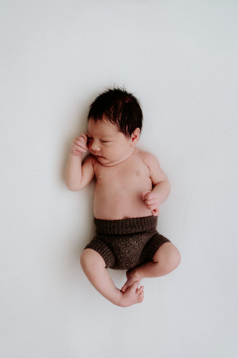 A sleeping newborn baby wearing brown knit shorts lies on a white background in a minimalist setting.