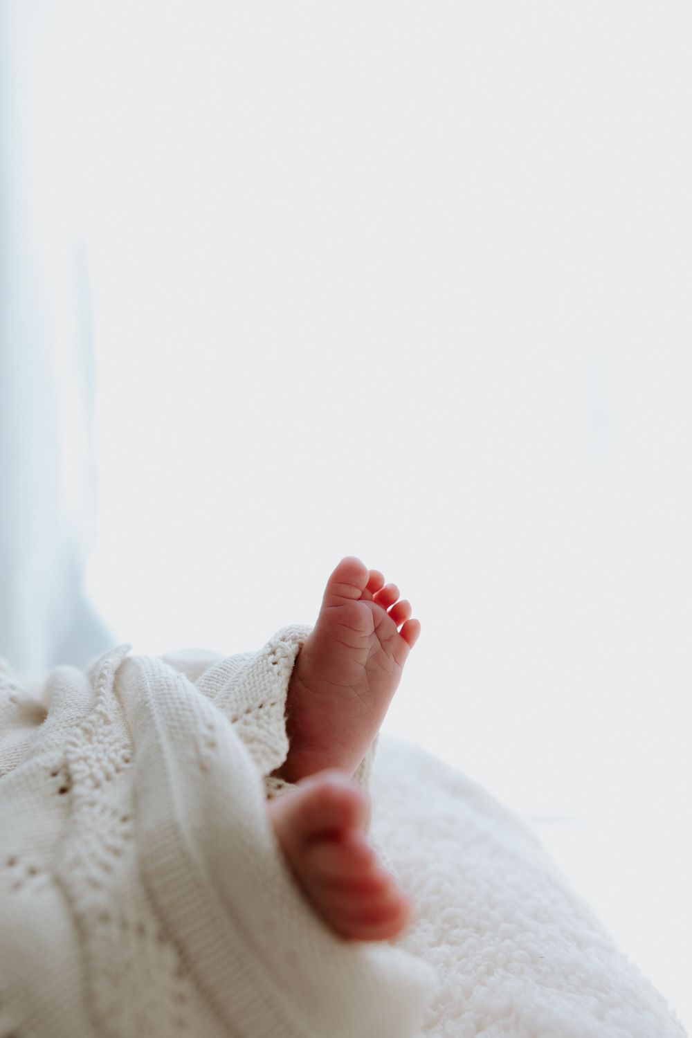 Tiny newborn baby feet peek out from beneath a soft white knitted blanket in a gentle natural light setting.