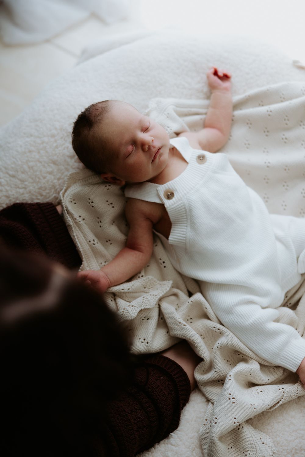A sleeping baby lies on soft white fabric, wearing a white eyelet dress against a light cream background.