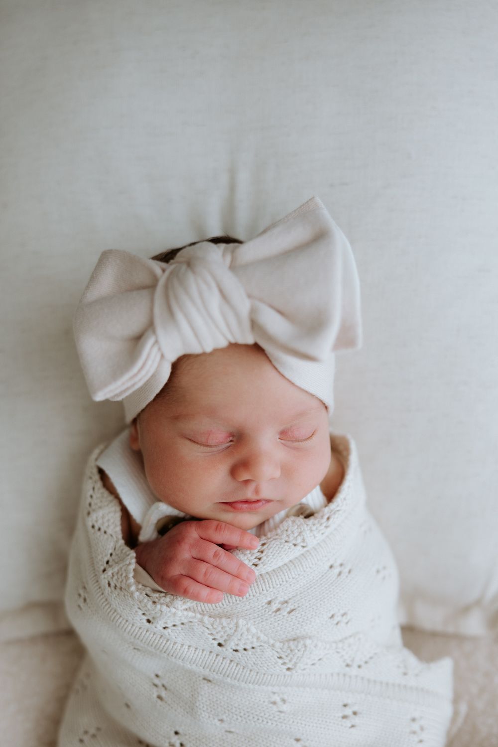 A sleeping newborn wrapped in a soft white blanket with a matching white bow headband rests peacefully.