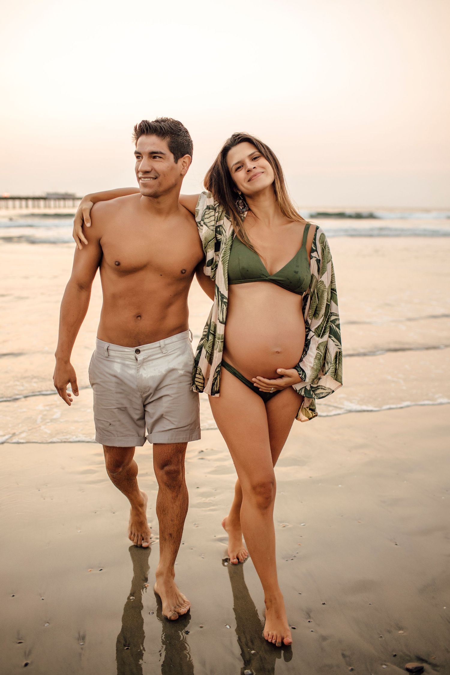 Expectant couple walking along beach at sunset wearing casual attire.