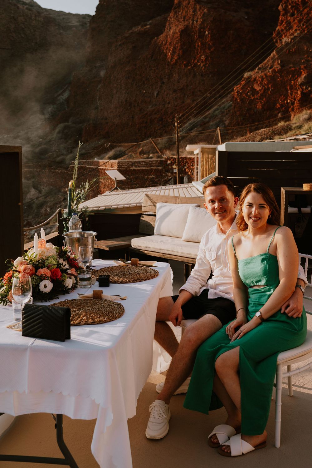 Romantic sunset dinner setup on a terrace overlooking dramatic canyon cliffs with floral centerpiece and table settings.