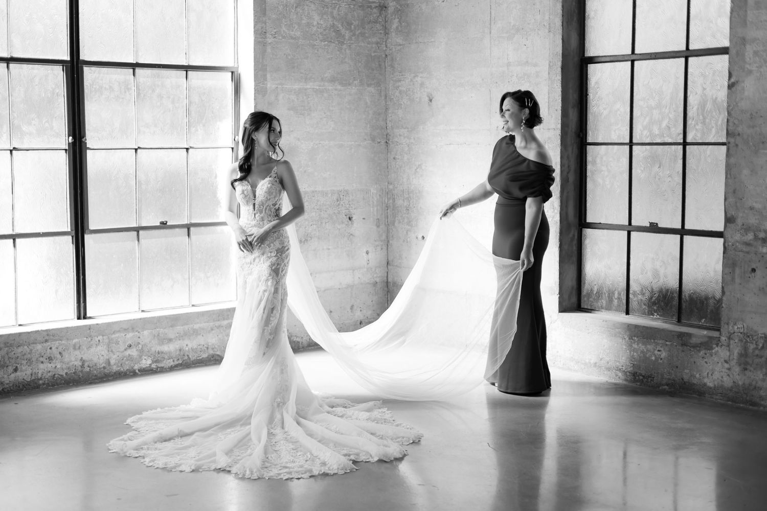 A bride in a lace gown has her wedding dress train adjusted in a bright industrial space.