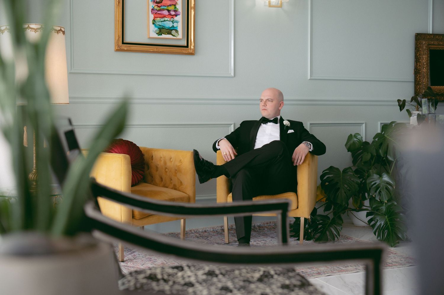 Groom in a black tuxedo relaxes on a golden velvet chair in an elegant room with houseplants.