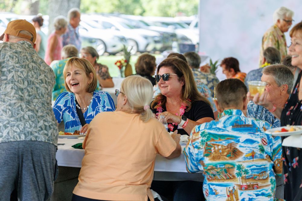 Group of smiling women enjoying a festive outdoor luncheon event, laughing and socializing at a decorated table.
