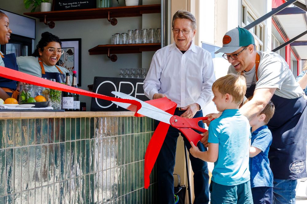 A red ribbon cutting ceremony takes place at a small business establishment with several people gathered around.