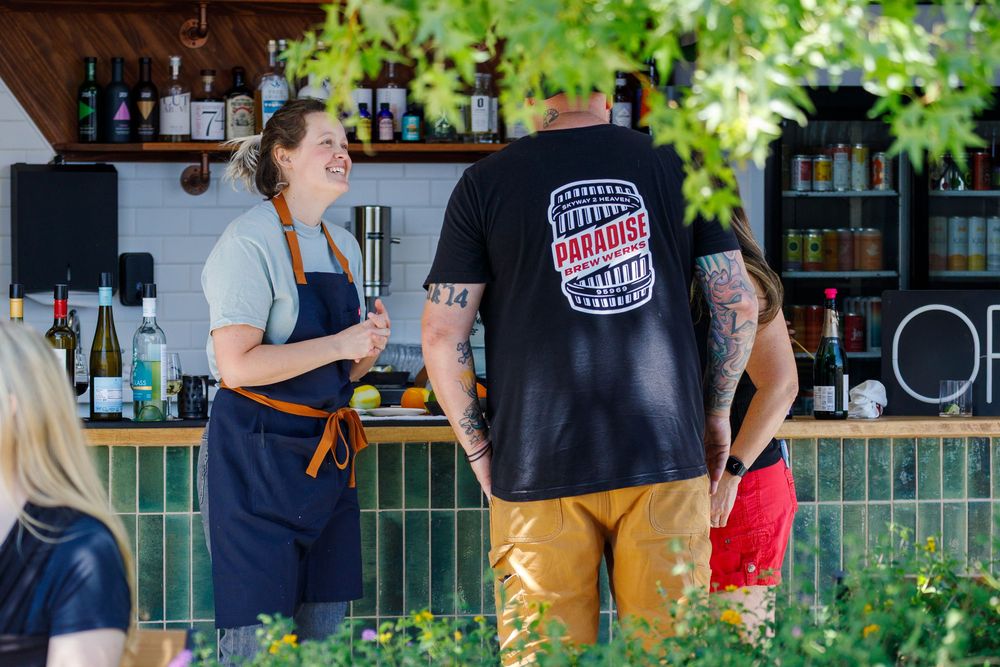 Customers order drinks at an outdoor bar with lush greenery and bottles on display in the background.