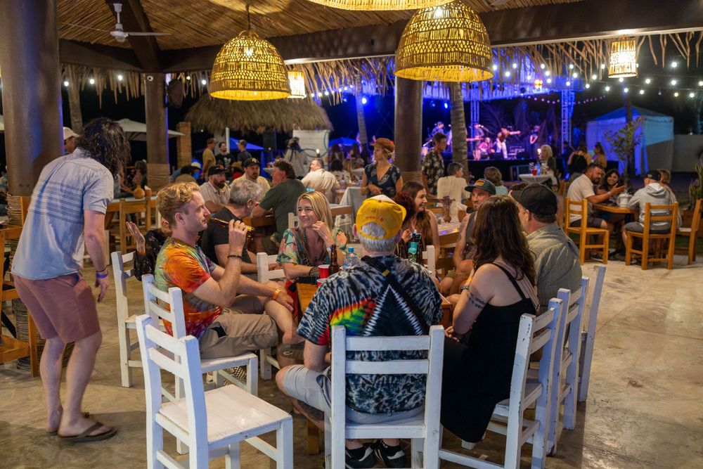 Busy restaurant scene with diners seated at white tables under woven pendant lamps.