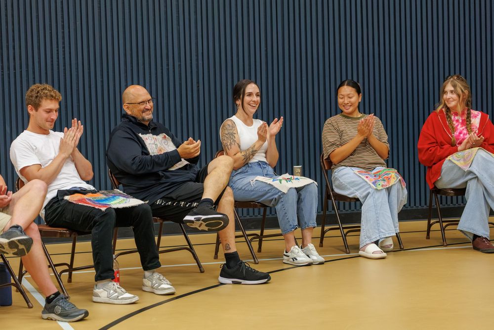 Five diverse young adults sit in chairs clapping and smiling in a gymnasium setting.