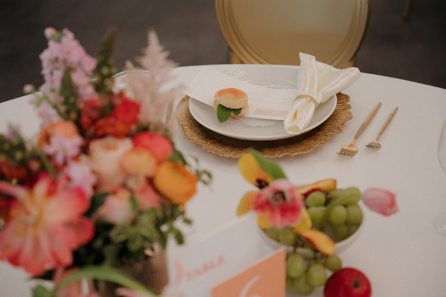 An elegant place setting with pink and coral flowers, gold charger plates, and fresh fruits on a white tablecloth.