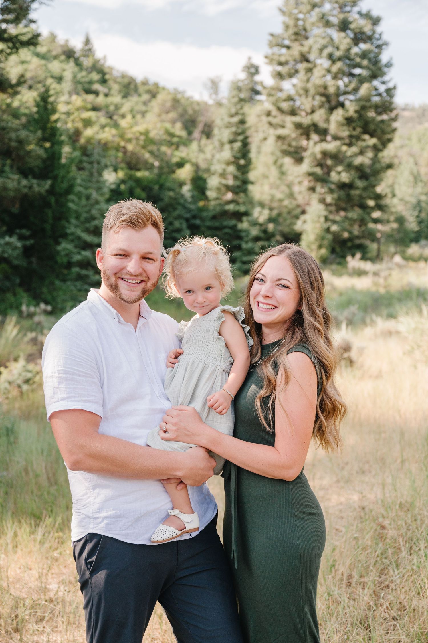 Happy family portrait in a scenic mountain meadow with evergreen trees in the background.