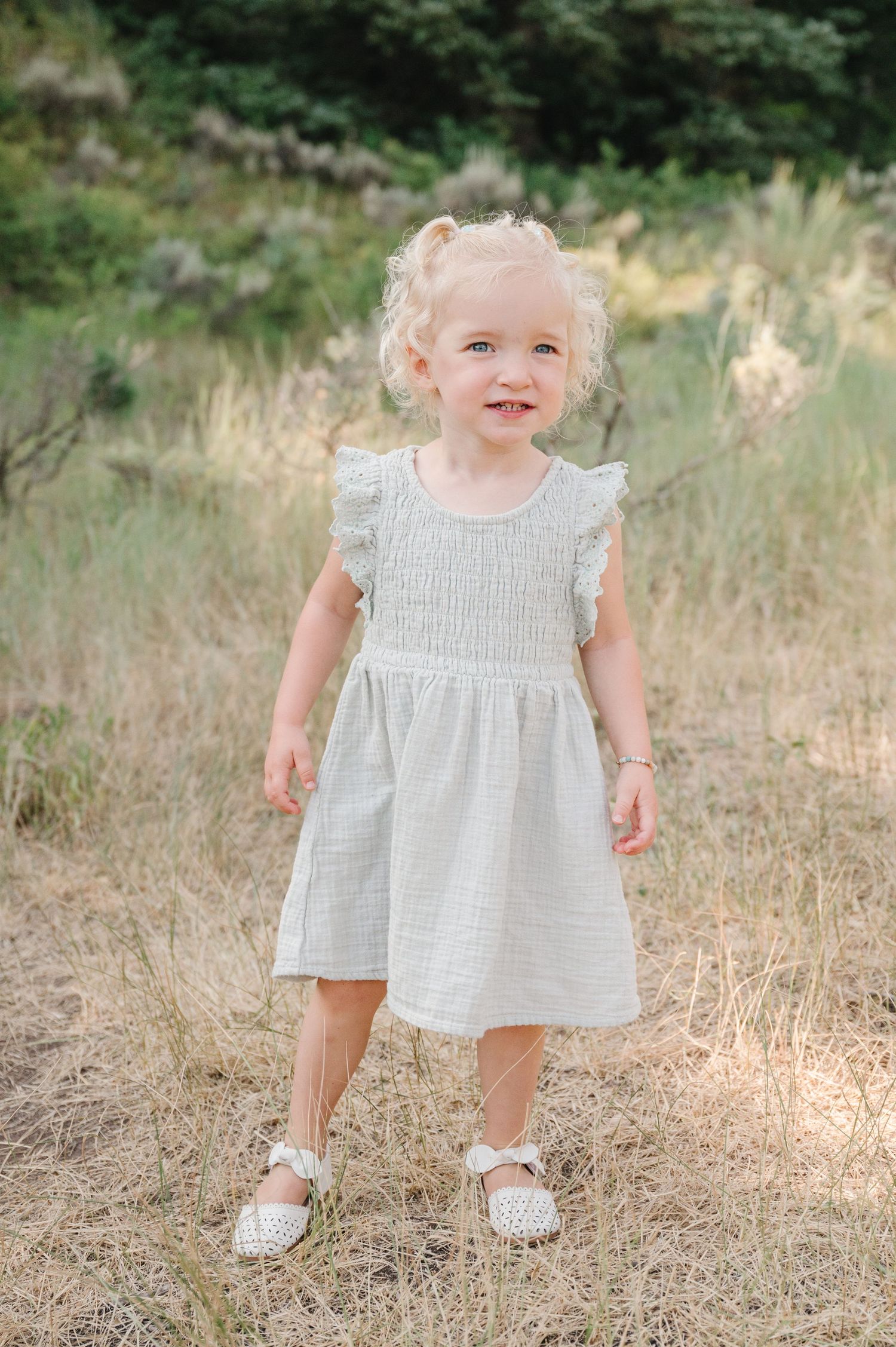 Little girl in white dress smiles while standing in mountain meadow.
