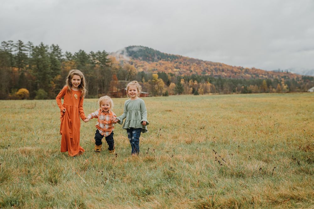 Three young siblings stand together in a vast autumn meadow with colorful fall foliage and mountains behind them.