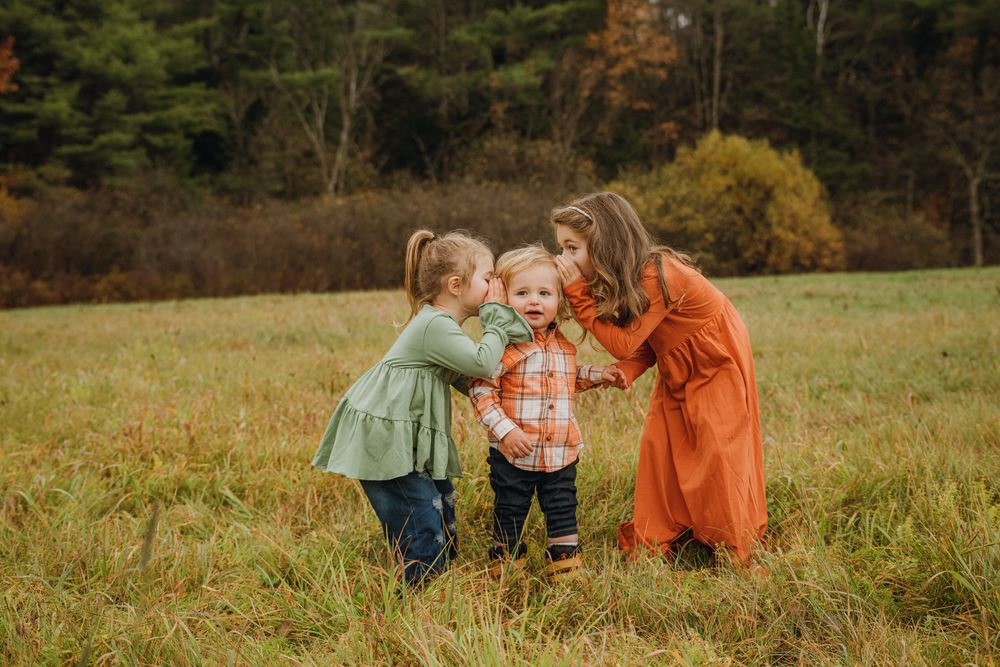 Two sisters lovingly kiss their baby sibling on the cheeks in a golden autumn field.