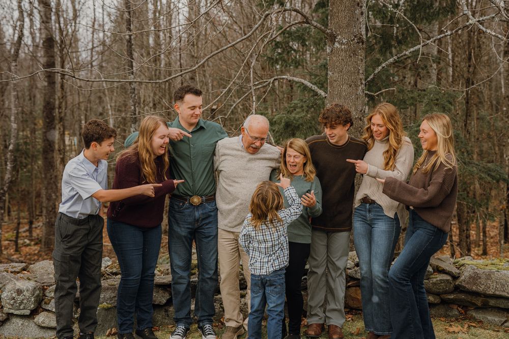 A multigenerational family gathers for an outdoor portrait in a wooded setting, sharing a warm embrace among bare trees.