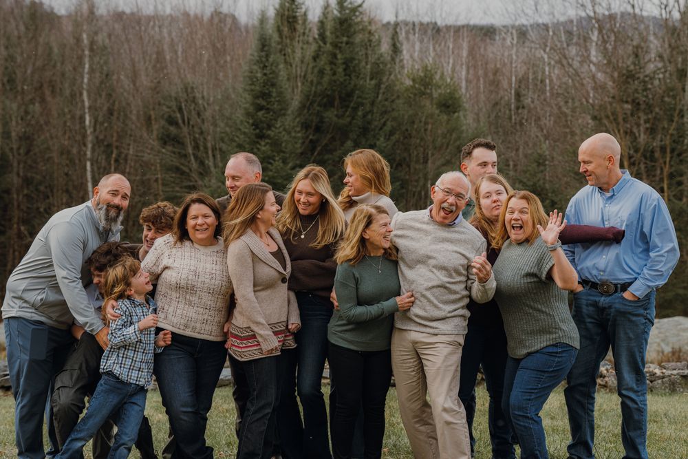 Large multigenerational family laughing together outdoors in a green field with autumn trees in the background.