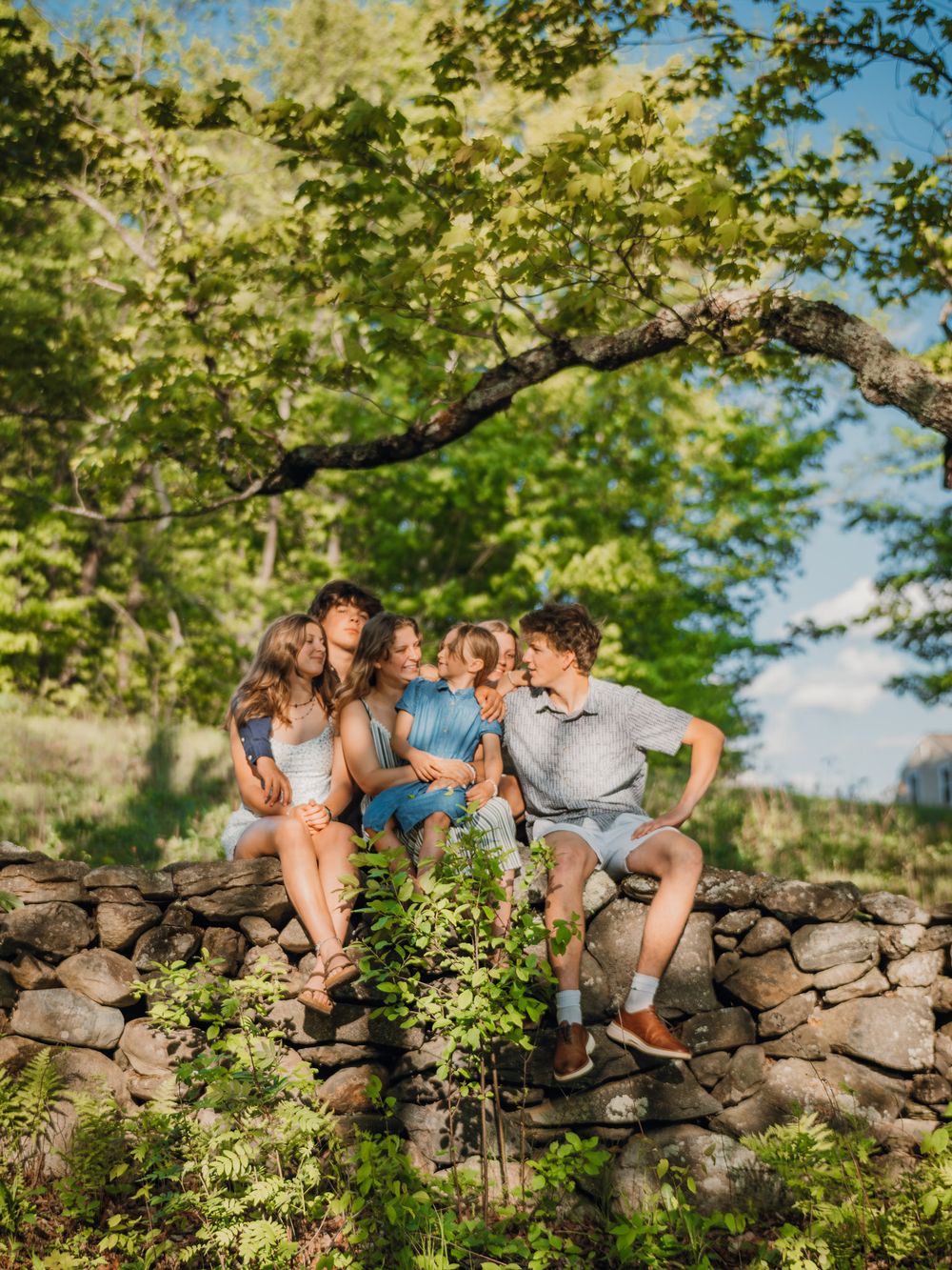 Family of four sitting on a rustic stone wall surrounded by lush green trees on a sunny day.