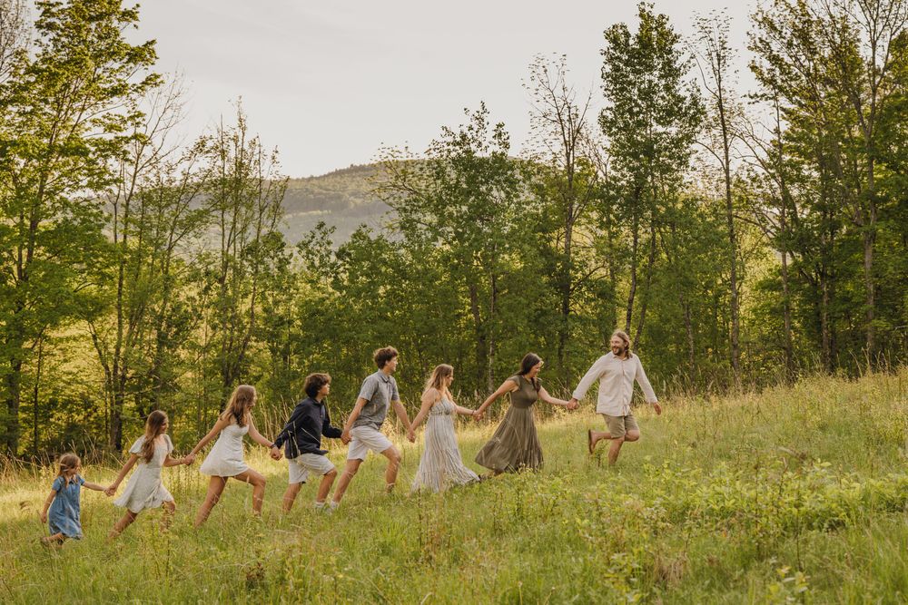 A joyful family holds hands running through a lush green meadow with tall trees and mountains in the background.