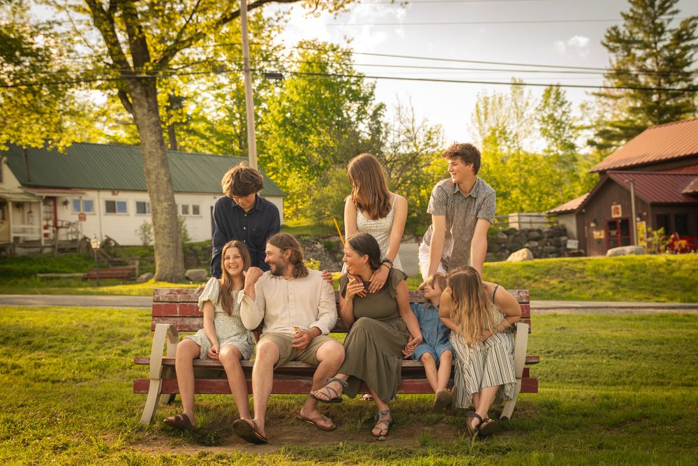Group of friends and family gathered on a park bench outdoors during golden hour in a lush green setting.