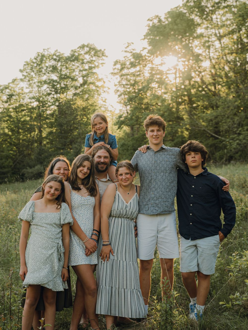 Large family of seven posing together in a sunlit meadow with trees in the background.
