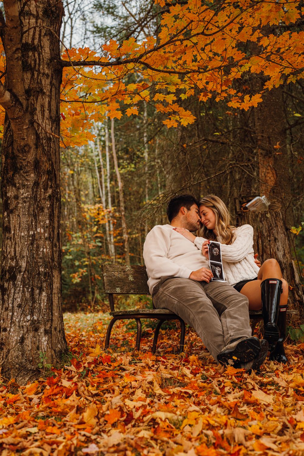Couple shares a romantic kiss on a bench surrounded by vibrant orange fall foliage while holding an ultrasound photo.