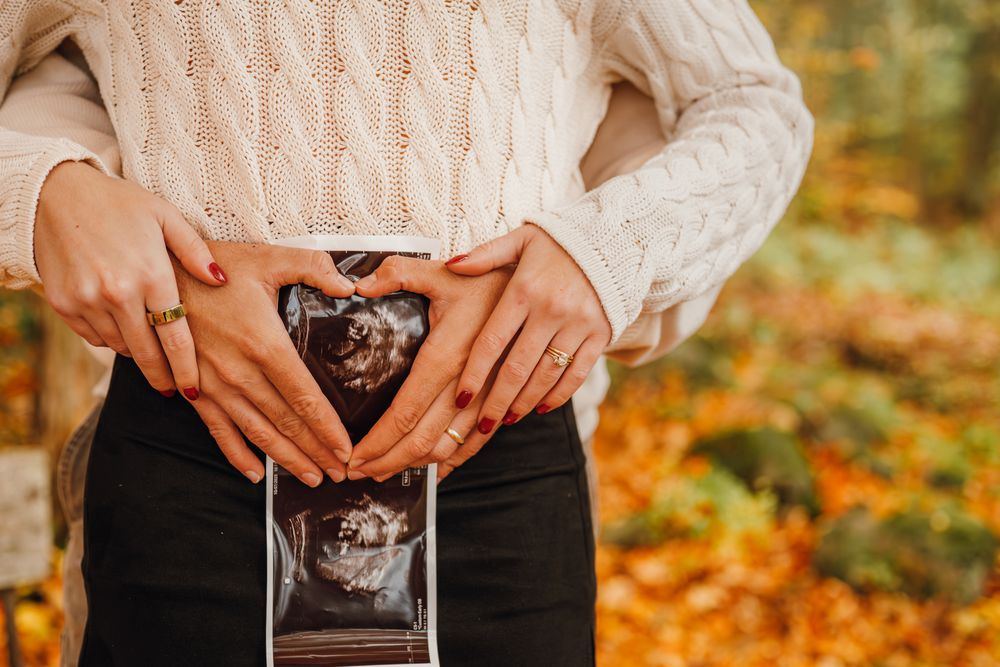 Couple forms a heart with their hands over an ultrasound photo against a colorful autumn background.