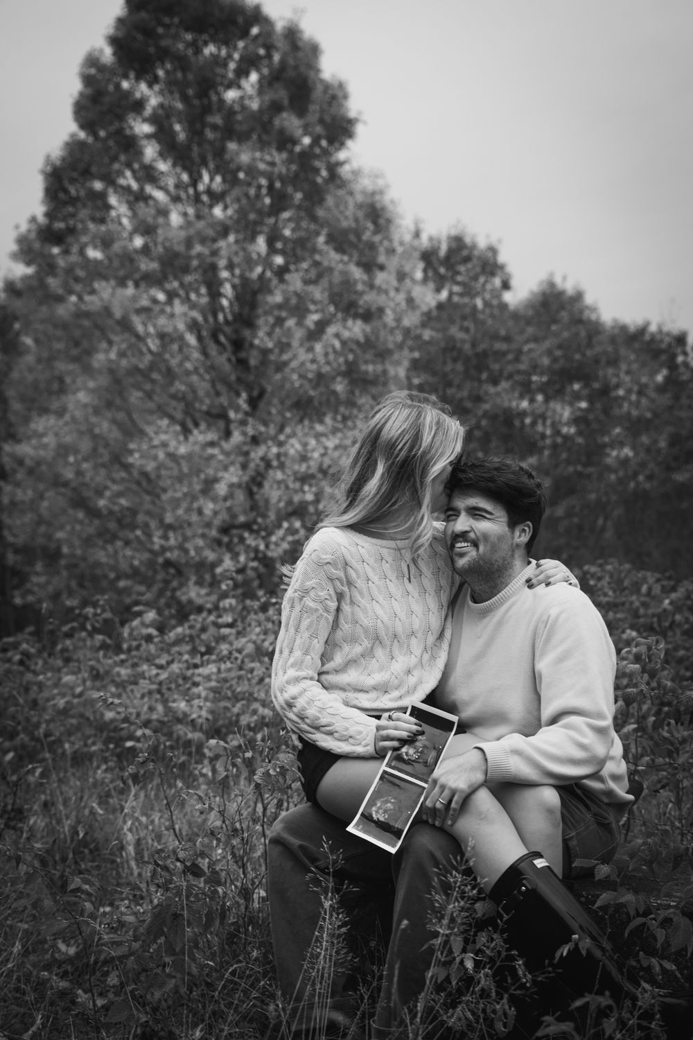Happy couple sits together in a field holding an ultrasound photo in a black and white pregnancy announcement.