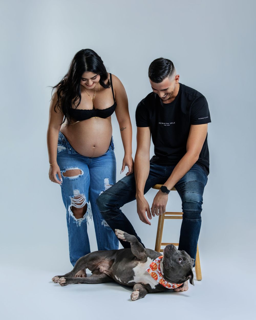 Pregnant couple in black tops and ripped jeans posing with a gray pit bull wearing a floral bandana in a studio.