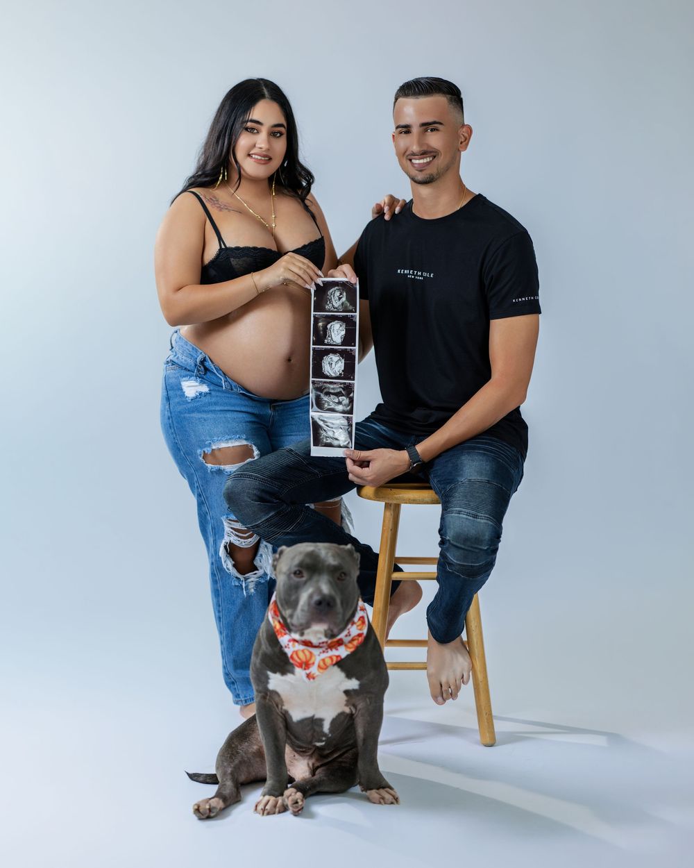 Smiling pregnant couple holding ultrasound photos with their gray pit bull dog wearing a bandana in a photo studio.