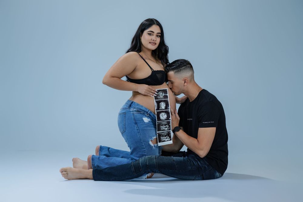 Couple sitting on studio floor, man kissing pregnant belly while holding ultrasound images, woman in black bralette.