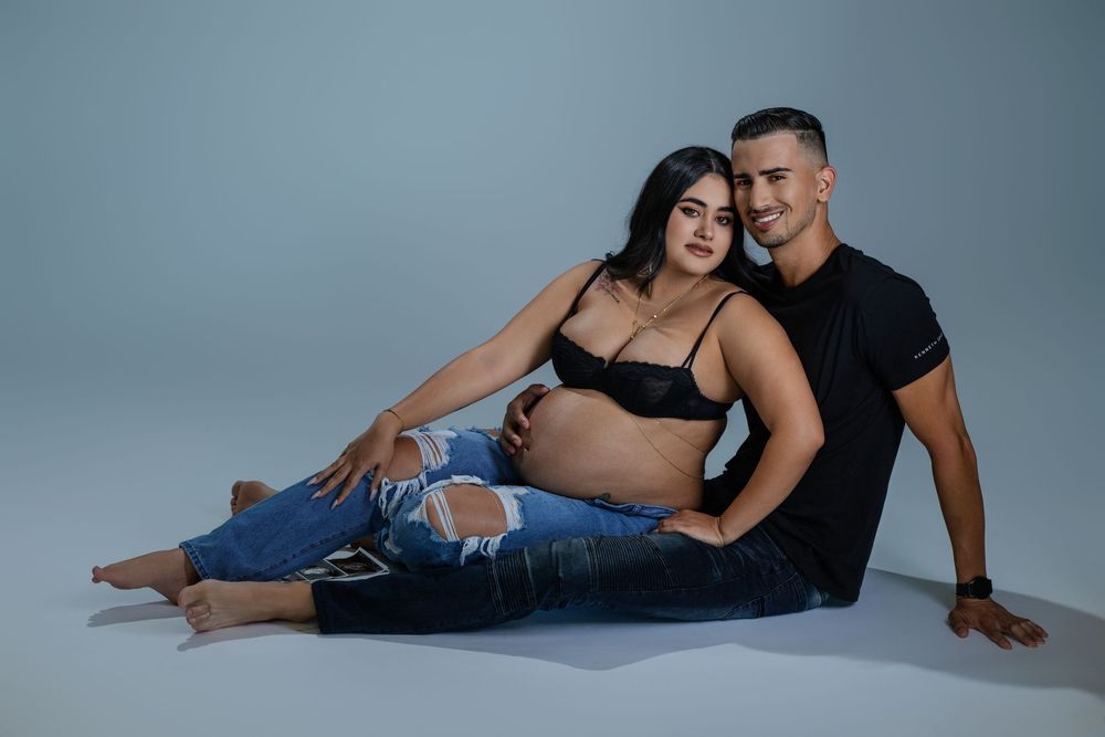 Pregnant couple sitting together on studio floor, woman in black bralette showing baby bump, man embracing her warmly.