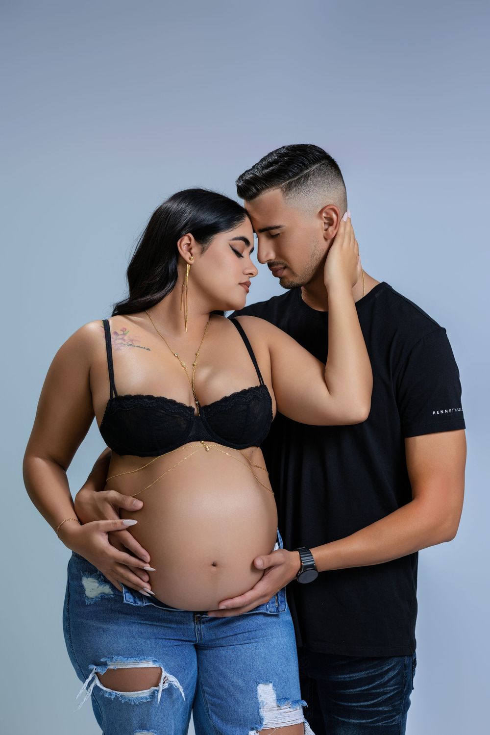 Couple with foreheads touching, man holding pregnant womans bare belly, both wearing black in a light blue studio.