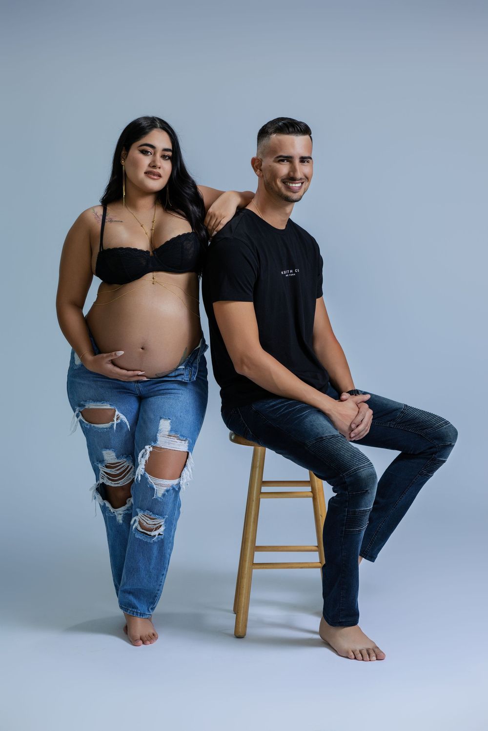 Smiling pregnant woman in black bralette and ripped jeans posing with partner on stool against light blue background.