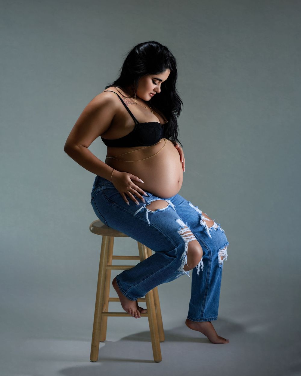 Pregnant woman in black bralette and ripped jeans sitting on wooden stool, cradling baby bump in a photo studio.