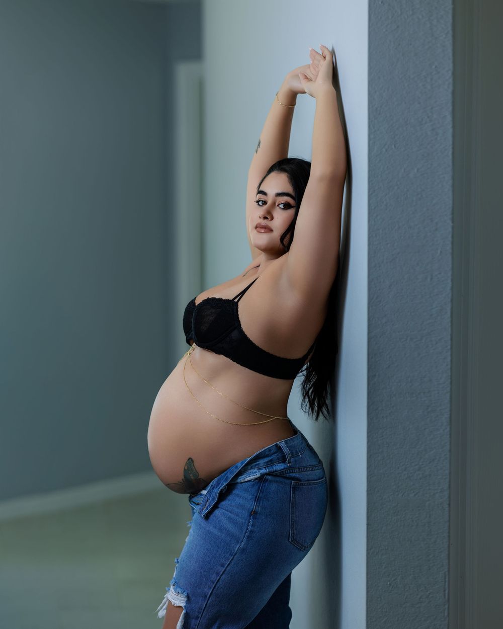 Pregnant woman in black bra and jeans posing against wall with arms raised in maternity photo shoot.