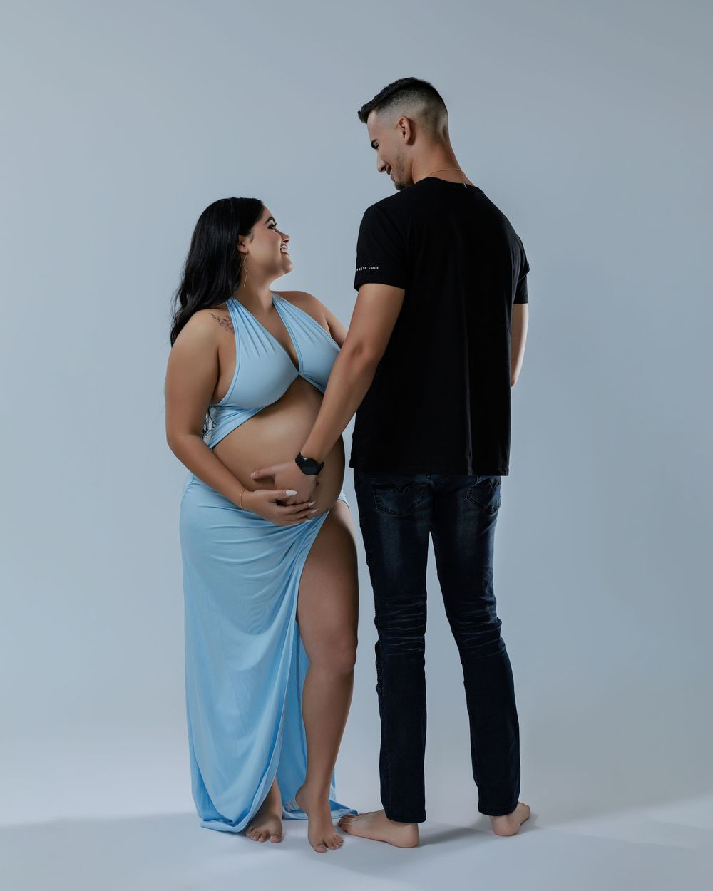 Laughing pregnant woman in light blue halter dress holding belly while facing her partner in a bright studio setting.