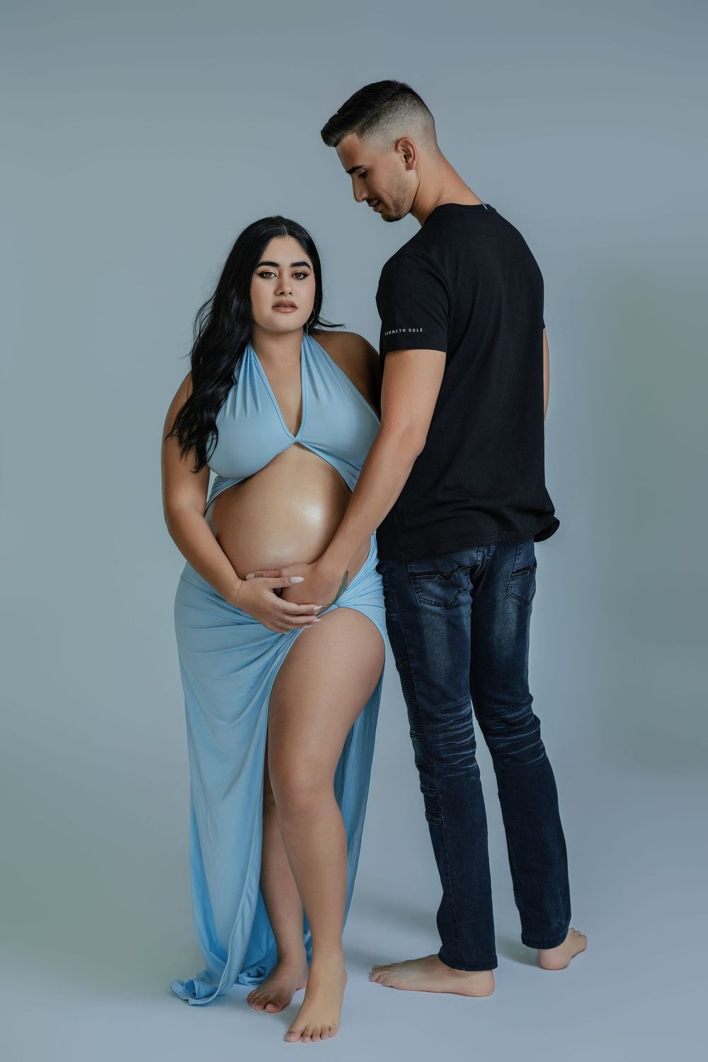 Couple expecting baby posing together in studio, woman in blue draped maternity gown, man in black shirt.
