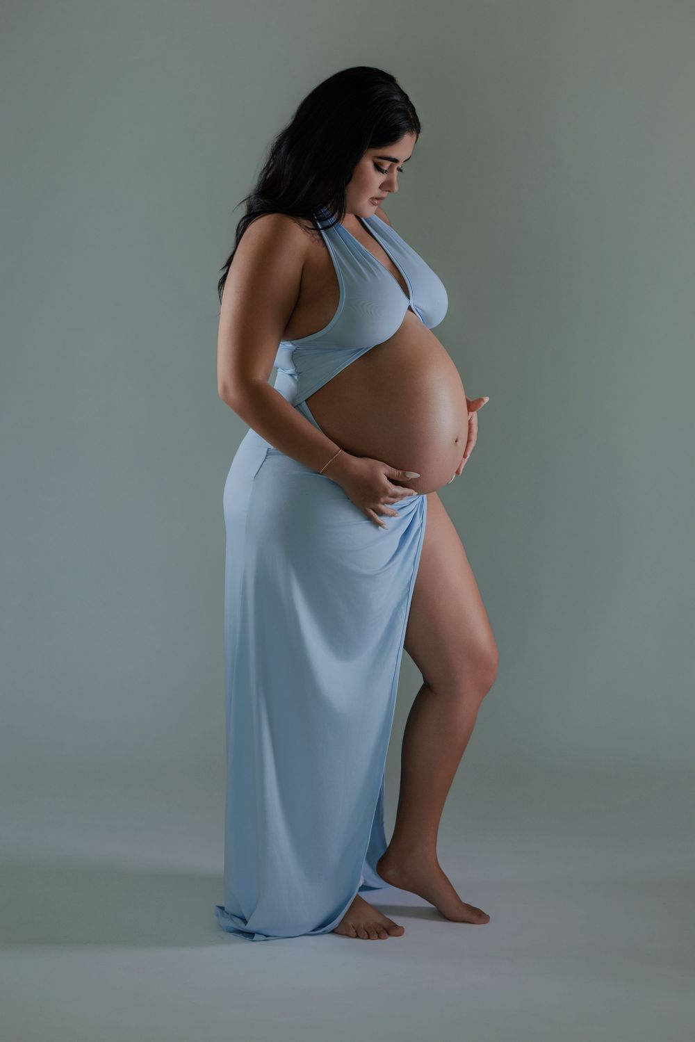 Pregnant woman in flowing light blue halter maternity gown holding baby bump in studio portrait.