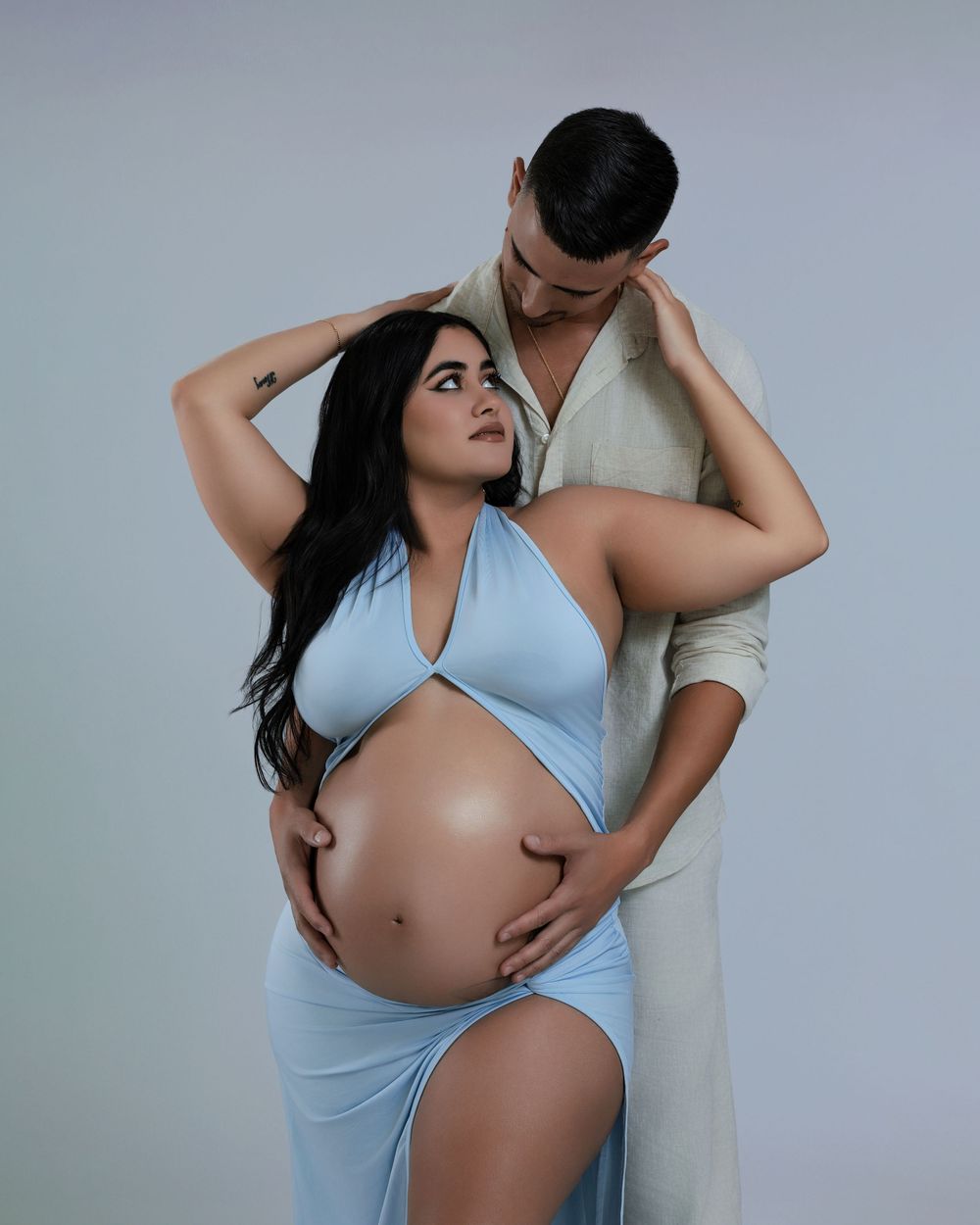 Expecting couple embracing in studio maternity shoot, woman in blue halter dress cradling pregnant belly.