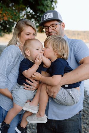 Loving family portrait with parents holding two young boys sharing a sweet kiss on a rocky beach.
