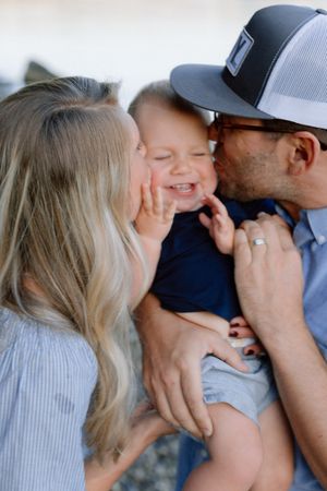 Parents simultaneously kissing laughing baby boy in navy outfit in a sweet family moment.