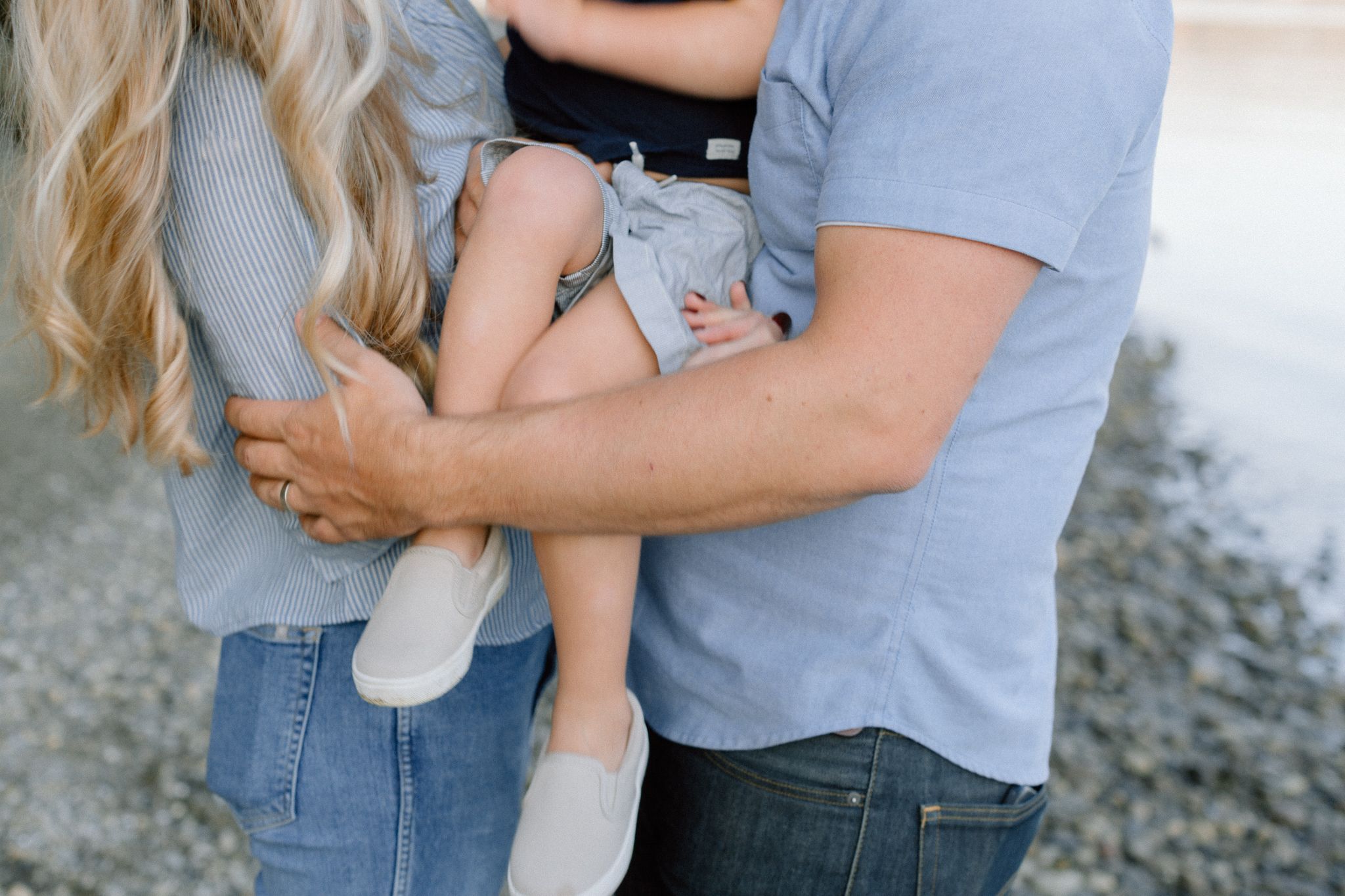 Close-up of parents in blue outfits holding young child with white sneakers outdoors.